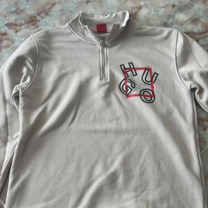 Beige half zip tröja från Hugo Boss - Säljer en beige långärmad tröja från Hugo Boss med half zip och hög krage. Framsidan har en stor HUGO-logga i svart och rött tryck. Tröjan är i mjuk bomull och har en clean, stilren look som passar perfekt till jeans eller joggers.