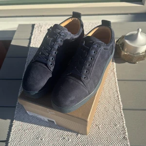 Mörkblå mocka sneakers från Louboutin - Snygga mörkblå sneakers från Christian Louboutin i lyxig mocka med klassisk röd sula och broderad logga baktill. Skorna har rund tå, snörning och diskreta detaljer som ger en clean och exklusiv vibe. Perfekta för dig som vill sticka ut med stil.