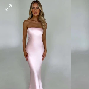 Ljusrosa fin klänning  - Superfin axelbandslös långklänning i ljusrosa satin-look. Klänningen har en figurnära passform, öppen rygg med snygg detalj och slät, glansig yta som ger en lyxig känsla. Perfekt för bal eller festliga tillfällen!