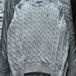 Grå kabelstickad tröja Polo Ralph Lauren - Knappt använd kabelstickad grå tröja från Polo Ralph Lauren med klassisk rund halsringning och broderad logga på bröstet. Tröjan är långärmad och har ribbade muddar vid ärmslut och nederkant. Perfekt för lager-på-lager och chill stil.  