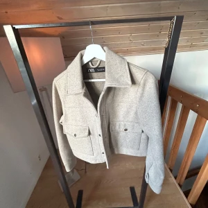 Beige overshirt från Zara - Snygg beige kappa från Zara med stora bröstfickor och knappar framtill. Jackan har en rak, croppad passform och är tillverkad i ett mjukt ullblandat material. 