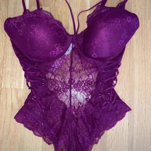 spetskorsett/body - Aldrig använd! Kupa: 85D. Magneta Lila färg korsett som är en body med detaljer, spets och push-up kupor. Sidorna har dekorativ snörning och tyget är genomskinligt med fina spetsdetaljer. Justerbara axelband och hyskknäppning bak. 