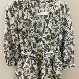 Vit och grön blommig klänning Zara - Superfin kort klänning från Zara i vitt med grönt blommönster. Klänningen har v-ringning, knappar framtill, trekvartsärm och ett bälte med rund spänne i midjan. Tillverkad i luftig bomull, perfekt för varma dagar. Ny 