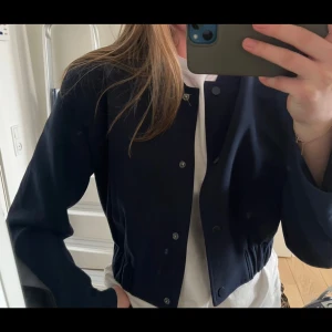 Mörkblå bomberjacka från Gina Tricot - Snygg mörkblå bomberjacka från Gina Tricot med croppad passform och tryckknappar framtill. Jackan har långa ärmar, elastisk midja och en stilren, minimalistisk look. Perfekt att slänga över en t-shirt för en chill vibe.