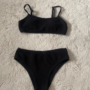 Svart ribbad bikini med raka band - Snygg svart bikini med ribbad struktur och raka axelband. Överdelen har enkel design med justerbara band och metallspänne i ryggen. Underdelen är klassisk med hög midja. Perfekt för stranden eller poolen, stilren och enkel att matcha. Vadderade kupor💕