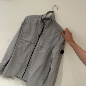 Grå vindjacka från C.P. Company  - Säljer en stilren grå vindjacka från C.P. Company med klassisk linsdetalj på ärmen. Jackan har två stora bröstfickor, dragkedja framtill och meshfoder på insidan. Perfekt för dig som gillar clean och funktionell streetwear.
