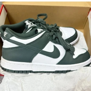 Nike Dunk Low vit och grön - Nike Dunk Low sneakers i vitt och mörkgrönt skinn. Klassisk siluett med rund tå, snörning och vit mellansula. Grön yttersula och detaljer, samt Nike-logga på hälen och tungan. Perfekta för dig som gillar streetstyle och vill sticka ut med en clean look.