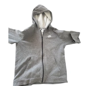 Grå hoodie från Nike med dragkedja - Grå hoodie från Nike med vit logga på bröstet. Tröjan har hel dragkedja, huva med mjukt fleecefoder och två stora fickor framtill. Perfekt för chill dagar eller när du vill ha en enkel och stilren look. (XL i barn storlek)