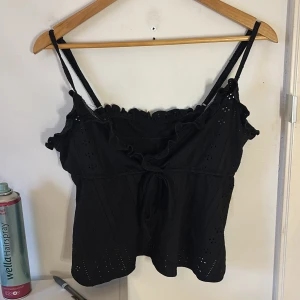 Svart linnetopp med volang och knyt - Supersöt svart linnetopp med smala axelband, volangdetaljer längs urringningen och knyt framtill. Toppen har broderade hålmönster som ger en luftig känsla och en feminin touch. Perfekt att styla med jeans eller kjol för en trendig look.