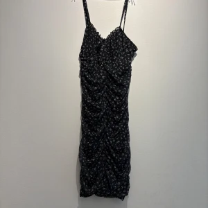 Svart blommig kortklänning DIVIDED - Supersöt svart kortklänning från DIVIDED med små blommor i vitt och rosa. Klänningen har smala axelband, rynkade detaljer framtill och en figurnära passform. Perfekt för dig som gillar en feminin och trendig stil.