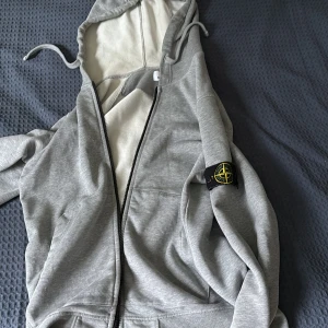 Grå hoodie från Stone Island - Snygg grå hoodie från Stone Island med dragkedja och klassisk logga på ärmen. Tröjan har huva med snörning och är tillverkad i mjuk bomull. Perfekt för dig som gillar streetwear och vill ha en stilren look.