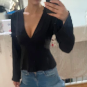 Svart omlottopp med rosett - Säljer en tröja med djup v-ringning och långa ärmar. Toppen har en snygg knytrosett i midjan som ger en feminin siluett. Perfekt att styla med jeans eller kjol för en trendig look. Fråga Gina