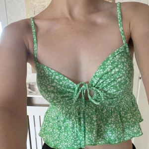 Grön blommig croppad linnetopp - Supersöt grön croppad linnetopp med vita småblommor och smala axelband. Toppen har en snygg knytning framtill och volangkant nedtill som ger en extra feminin touch..