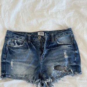 Blå jeansshorts från Vero Moda - Snygga blå jeansshorts från Vero Moda med slitna detaljer och fransig nederkant. Klassisk femficksmodell med knapp och dragkedja framtill. Perfekta för sommardagar när du vill ha en avslappnad och trendig look.