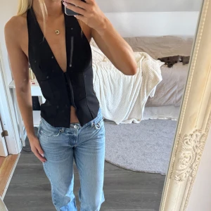 Svart omlottväst med djup v-ringning - Säljer en svart väst med djup v-ringning och omlottkänsla. Västen är ärmlös, har öppen rygg och stängs framtill. Sitter tight och har en stilren, minimalistisk look som passar perfekt till jeans eller kjol.