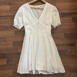 Vit kortklänning med broderade prickar - Supersöt vit kortklänning från Chelsea med broderade prickar, v-ringning och knappar framtill. Klänningen har korta puffärmar med resår och markerad midja. Perfekt för sommarens alla tillfällen!