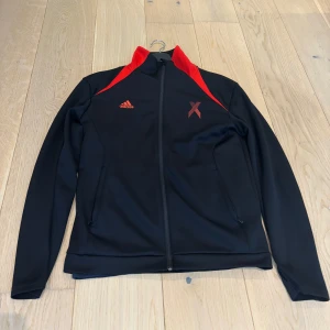Svart Adidas träningsjacka med röd detalj - Svart träningsjacka från Adidas med röda detaljer på axlarna och stort rött X på ryggen. Säljs inte någon stans längre och är förmodligen den ända ni kommer att hitta S/M 