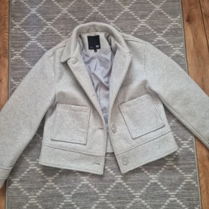 Ljusgrå kappa - Stilren ljusgrå kappa från lager 157 med klassisk krage, stora framfickor och knäppning framtill. Jackan har en rak passform och är fodrad för extra komfort. Perfekt för dig som gillar en clean och modern look. Nypris 400kr och är slutsåld nästan överallt