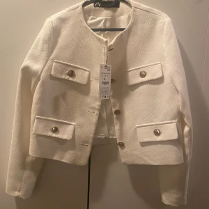 Vit kavajjacka från Zara, storlek S - Stilren vit kavajjacka från Zara i strukturerat tyg med fyra fickor framtill och snygga guldfärgade knappar. Jackan har rak passform, rund halsringning och är helfodrad. Perfekt för dig som vill ha en chic och trendig look.