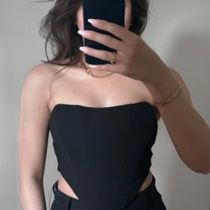 Svart korsett-topp med dragkedja - Säljer en svart axelbandslös korsett-topp med markerad midja och snygga utskärningar vid sidorna. Toppen har en dragkedja bak och är croppad för en edgy look. Perfekt att styla med höga byxor eller kjol.