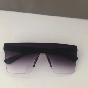 Balenciaga svarta shield-solglasögon - Snygga shield-solglasögon från Balenciaga med svart båge och stora, rektangulära linser i mörkgrå toning. Glasögonen har en futuristisk vibe med hel front och raka linjer. Perfekta för dig som vill sticka ut och köra på en edgy stil.
