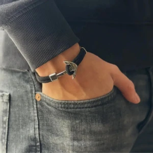 Svart armband med ankardetalj - Stilrent svart armband med en cool silverfärgad ankare som lås. Bandet är gjort av ett slitstarkt material och passar perfekt till en avslappnad look. Ankaret ger en nautisk vibe och armbandet är lätt att justera för bästa passform.