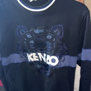 Svart Kenzo LIMITED tröja - Svart långärmad tröja från Kenzo med blåa detaljer och ikonisk tigerbrodyr framtill. Tröjan har en blå rand över bröstet och ryggen samt ribbad krage med vita och blå ränder. Perfekt för dig som gillar streetstyle och vill sticka ut.