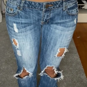 Blå slitna bootcut jeans - Säljer ett par blå bootcut jeans med slitningar och stora hål vid knäna. Jeansen har klassisk femficksdesign och är tillverkade i denim. Perfekta för en avslappnad och trendig look.