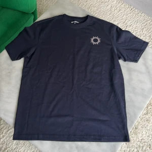 Mörkblå t-shirt från New Garments XS - Mörkblå t-shirt i 100% bomull från New Garments. Framtill finns en tryckt solsymbol och på ryggen står texten 'NOT THERE YET "It is my last year being broke."' i vitt. Klassisk passform med rund hals och korta ärmar. Perfekt för dig som vill ha en fet t-shirt till sommaren. Har aldrig använt storlek XS men sitter som S. 
