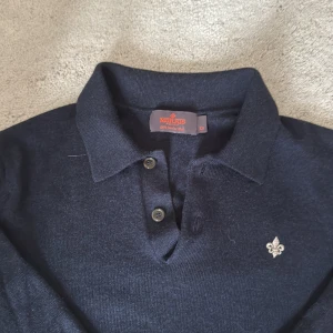 Mörkblå merinoull pikétröja Morris - Snygg mörkblå långärmad pikétröja från Morris i 100% merinoull. Klassisk krage, två knappar och diskret broderad logga på bröstet. Storlek xs