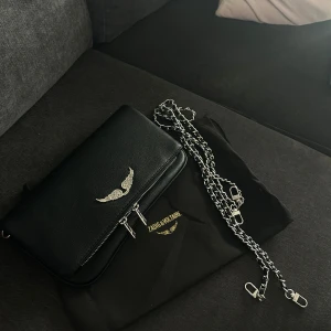 Svart axelväska Zadig & Voltaire - Snygg svart axelväska från Zadig & Voltaire i skinn med silverfärgad kedjerem och coolt vingemblem framtill. Väskan har dubbla dragkedjor och stilrena detaljer i silver. Perfekt storlek för det viktigaste och ger en edgy vibe till din outfit.
