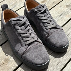 Grå mockasneakers från Louboutin - Snygga grå sneakers i mocka från Louboutin med klassisk röd sula. Skorna har rund tå, platt sula och breda grå skosnören. Insidan är i beige skinn och designen är stilren och lyxig, perfekt för dig som gillar exklusiva detaljer.