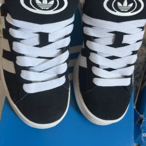 Adidas Campus sneakers svart & grå mocka - Två par Adidas Campus sneakers, ett par i svart mocka med vita detaljer och ett par i grå mocka med vita ränder. Båda har klassiska tre ränder på sidan, vit sula och breda vita skosnören. Perfekt för dig som gillar retrostil och streetwear. 