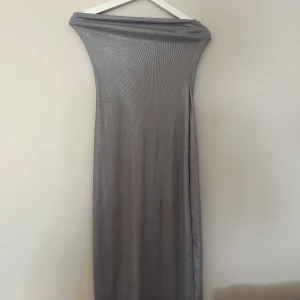 Silver axelbandslös långklänning H&M - Snygg axelbandslös långklänning från H&M i glansigt silverfärgat tyg. Klänningen är ribbad och har en figurnära passform som ger en cool och modern look. Perfekt för dig som vill sticka ut med en stilren och trendig vibe.