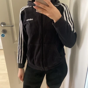 Svart Adidas hoodie med dragkedja - Svart hoodie från Adidas med klassiska vita ränder längs ärmarna och vit logga på bröstet. Jackan har huva, dragkedja framtill och två fickor. Perfekt för en sportig och chill stil.