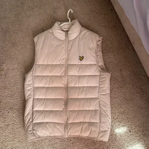 Beige dunväst från Lyle & Scott - Snygg beige dunväst från Lyle & Scott med klassisk gul logga på bröstet. Västen har quiltad design, hög krage och dragkedja framtill. Perfekt att slänga över en hoodie eller tröja för extra värme. Enkel och stilren look som funkar till det mesta.