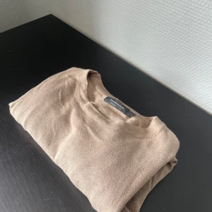 Beige stickad tröja Dressmann - En stilren beige stickad tröja från Dressmann. Tröjan har en klassisk rund halsringning och är tillverkad i mjukt material som passar perfekt för lager-på-lager. Enkel att matcha med jeans eller chinos för en clean look.