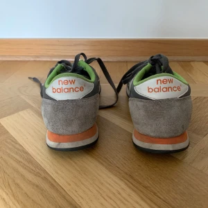 Grå New Balance sneakers med gröna detaljer - Snygga New Balance sneakers i grå mocka och textil med vita och orange detaljer. Skorna har grönt foderkant, klassisk N-logga på sidan och snörning. 