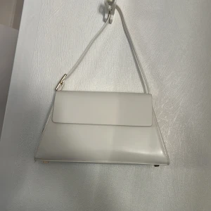 Vit axelväska med guldiga detaljer - Prislappen kvar, aldrig använd. Snygg vit axelväska i slät skinnimitation med stilren, triangulär form och guldfärgade metalldetaljer. Väskan har ett minimalistiskt uttryck och ett smalt axelband som ger en clean look. Perfekt för dig som gillar trendiga accessoarer med modern känsla.