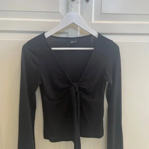 Svart knytblus från Gina Tricot - Svart långärmad topp från Gina Tricot med djup v-ringning och snygg knytning framtill. Toppens stilrena design gör den lätt att matcha och den har en figurnära passform. Perfekt för dig som gillar trendiga och enkla plagg.