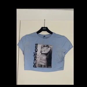 Blå croppad topp Ariana Grande - Croppad ljusblå topp från Ariana Grande merch med svart tryck 'positions' och ett stort svartvitt foto framtill. T-shirten har korta ärmar och rund halsringning. 