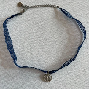 Blå spetschoker med peace-hänge - Chokerhalsband med blå spets och ett runt peace-märke i silverfärg, dekorerat med små glittrande stenar. Justerbart spänne i metall. Perfekt för att ge din outfit en cool och unik touch.