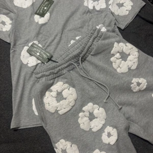 Grå t-shirt från Denim Tears med tryck - Grå t-shirt från Denim Tears med stora vita blommiga tryck över hela tröjan. Klassisk rund halsringning och korta ärmar. Mjuk bomullskänsla och riktigt chill vibe, perfekt för dig som gillar streetwear med unik design.