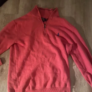 Korallfärgad tröja Polo Ralph Lauren - Snygg korallfärgad tröja från Polo Ralph Lauren i mjuk pima bomull. Tröjan har half zip-dragkedja, lång ärm och klassisk broderad logga på bröstet. Perfekt för dig som gillar stilrena och bekväma plagg.