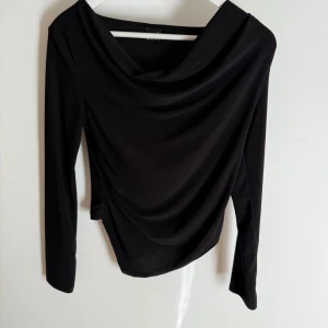 Svart draperad långärmad topp - Snygg svart långärmad topp med draperad halsringning och smal passform. Tyget faller mjukt och ger en elegant look. Perfekt att styla med jeans eller kjol för en trendig outfit.