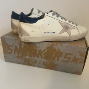 Golden Goose Superstar sneakers vit/silver - Golden Goose Superstar sneakers i vitt skinn med grå mockastjärna på sidan, grå snörning och mörkblå häl. Snygg vintage-look på sulan och detaljer i silver. Perfekta för dig som gillar streetstyle och unika detaljer.