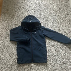 Vindjacka  - Snygg mörkblå vindjacka från Sail Racing med huva och hel dragkedja. Jackan har två sidofickor med dragkedja och diskret logga på bröstet samt en metallbadge på ärmen. Perfekt för blåsiga dagar och sportig stil. Priset kan diskuteras 