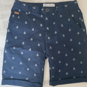 Mörkblå shorts med ankare från Zara - Snygga mörkblå shorts från Zara med vita ankare som mönster. Klassisk passform med fickor fram och bak, bälteshällor och knappstängning. Perfekta för sommardagar och har en stilren look med marina vibbar. Tillverkade i mjuk bomull för skön känsla.