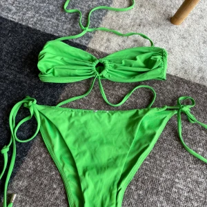 Grön bikini med knyt och ringdetalj - Använd TVÅ gånger!! Snygg grön bikini med bandeau-topp som har en rund ringdetalj i mitten och knytband både i nacken och ryggen. Bikinitrosan har höga ben och knyt i sidorna. Perfekt för stranden eller poolen, och färgen poppar verkligen i solen! 