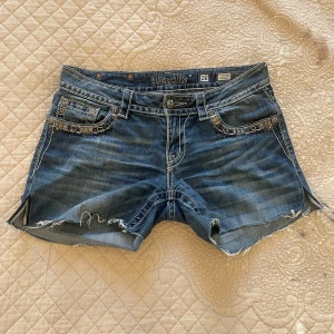 Miss Me jeansshorts med detaljer - Snygga blå jeansshorts från Miss Me med slitna kanter och coola broderade detaljer på fickorna. Shortsen har låg midja, dekorativa nitar och dragkedja. Perfekta för sommaren när du vill sticka ut lite extra.